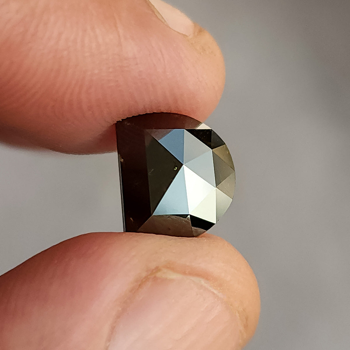 Tilted side-angle view of a D-shaped, dark brown gemstone held between fingers, highlighting the cut pattern on its curved edge.