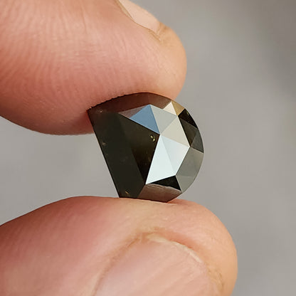 Hand-held D-shaped gemstone pinched between two fingers, showing faceted edges and deep brown color under bright lighting.