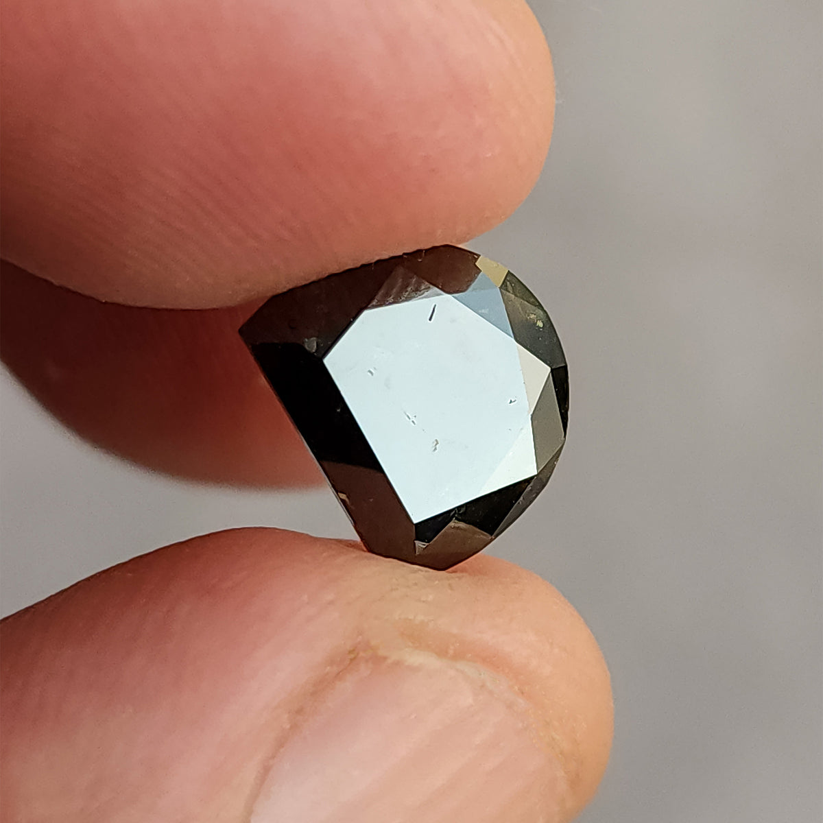 Dark, D-shaped cut gemstone gripped between thumb and forefinger, with its large top facet fully reflecting a bright, white light.