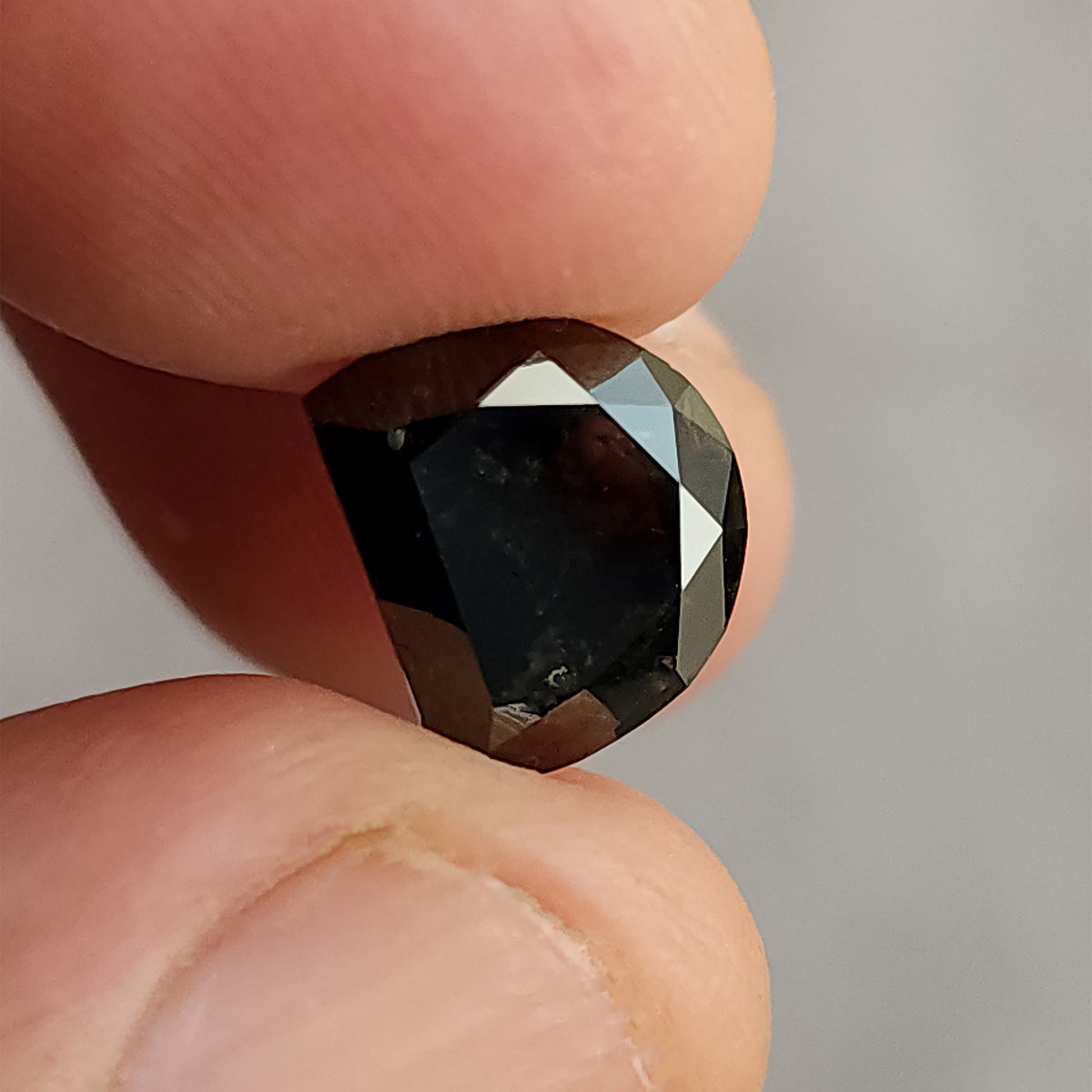 Very dark, D-shaped faceted gemstone pinched between fingers, angled to show the contrasting light reflections on its edge facets.
