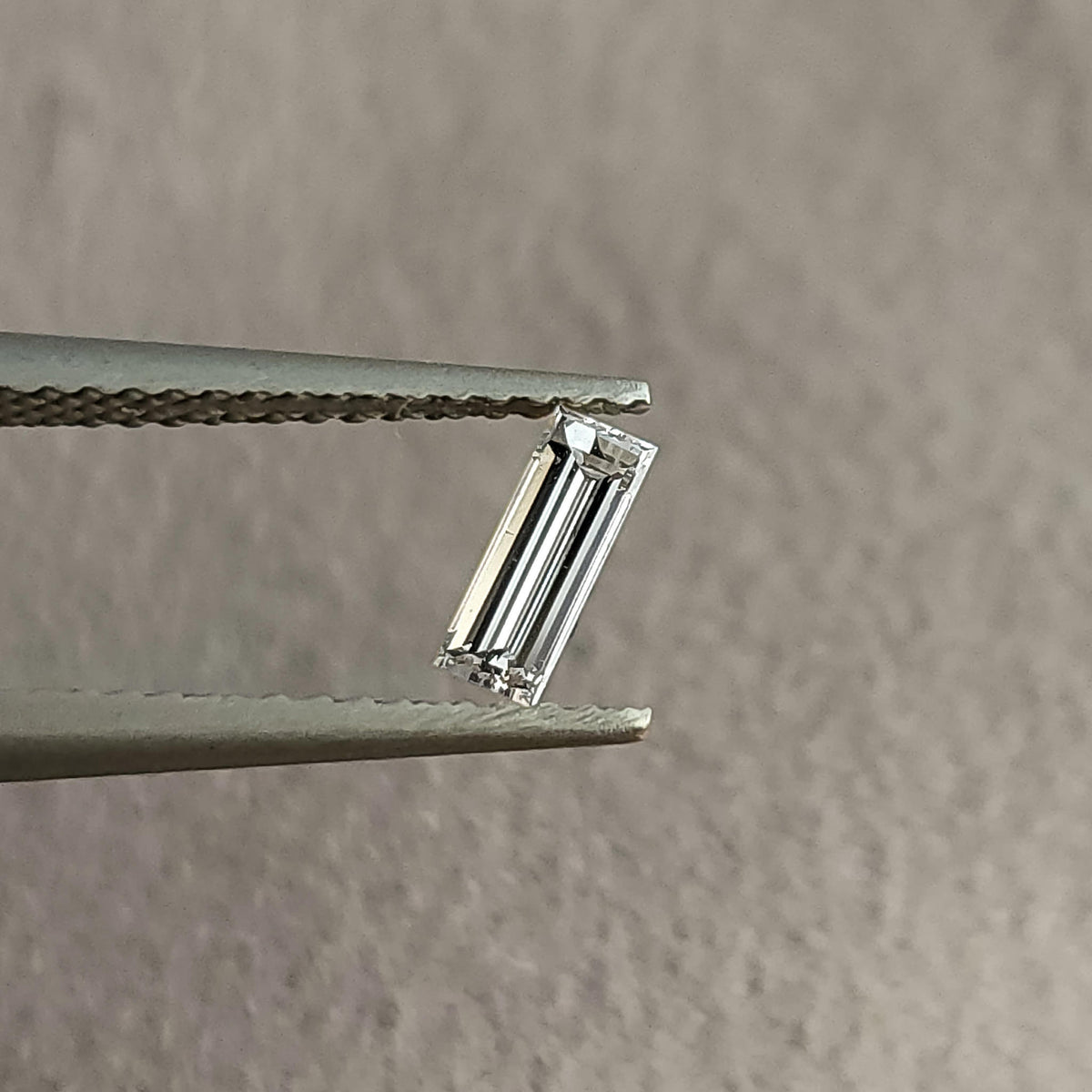 Face-up baguette-cut diamond held in tweezers against a neutral gray background, showing its long rectangular outline and parallel facets.