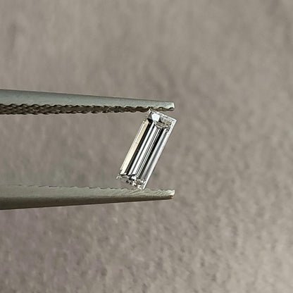 Face-up baguette-cut diamond held in tweezers against a neutral gray background, showing its long rectangular outline and parallel facets.