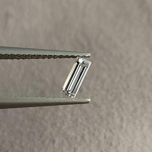 Face-up baguette-cut diamond held in tweezers against a neutral gray background, showing its long rectangular outline and parallel facets.