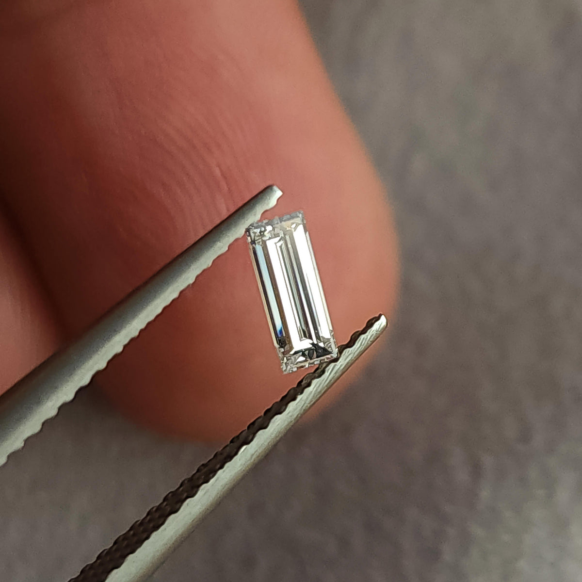 Face-up baguette diamond in tweezers with a fingertip behind it, giving scale for the narrow rectangular stone.