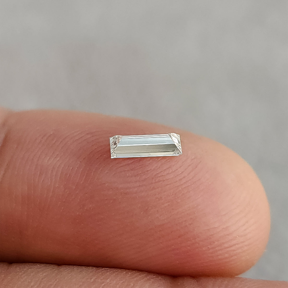Baguette diamond on a fingertip from a shallow angle, showing pale reflections across the flat-looking center.