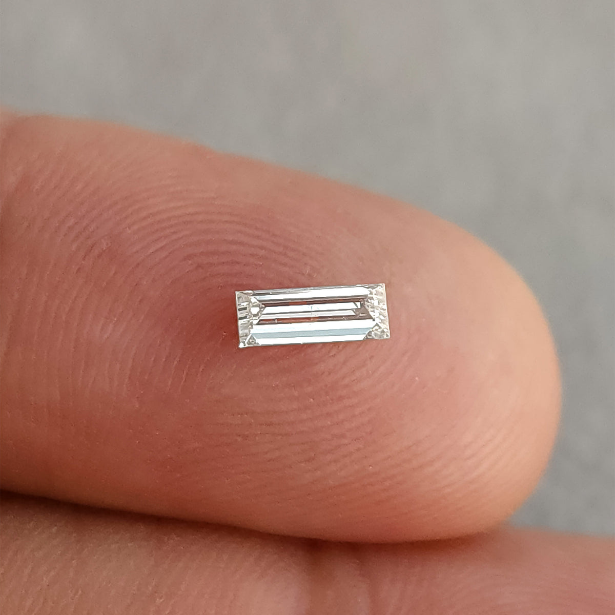 Alternate face-up fingertip view of the baguette diamond, showing bright parallel reflections.