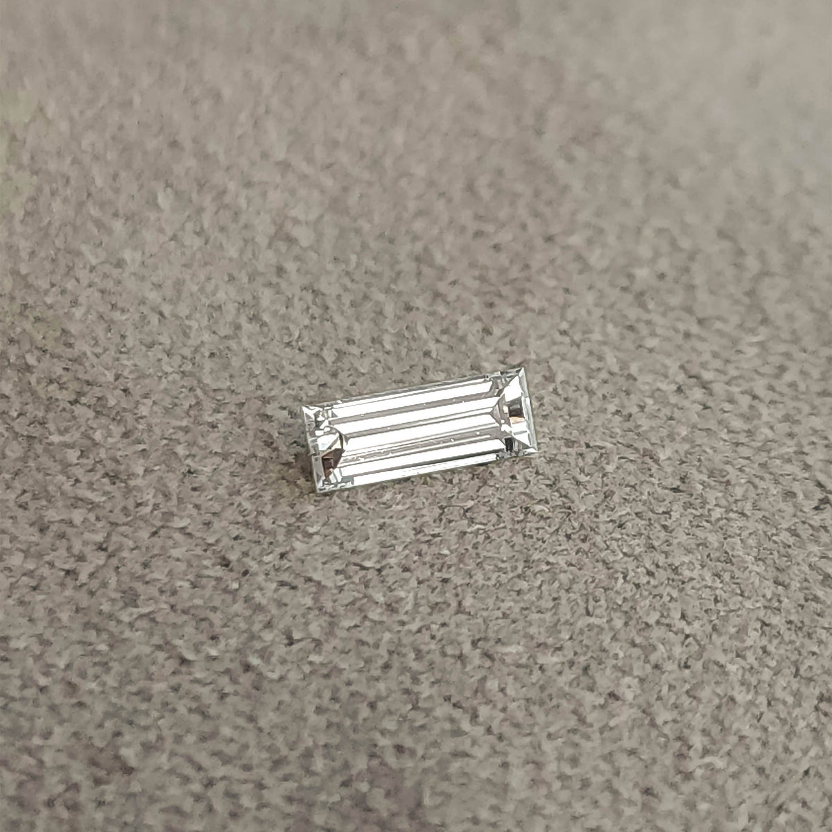 Baguette diamond lying face-up on a gray textured surface, showing its slim rectangular outline.