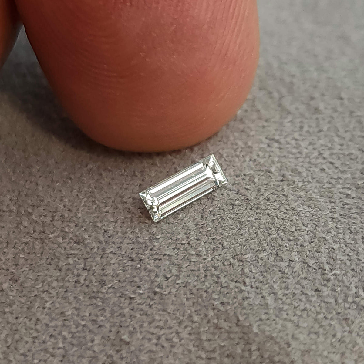 Baguette diamond on a gray surface beside a fingertip, providing a close scale comparison.