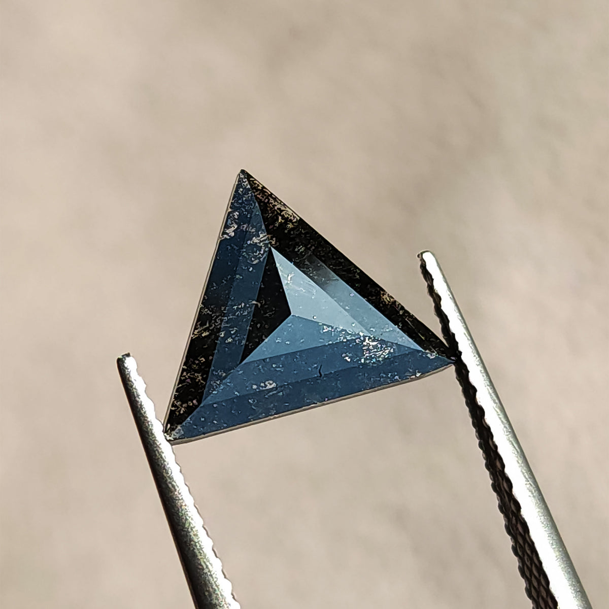 Face-up view of a dark triangular rose cut gemstone held vertically in tweezers.