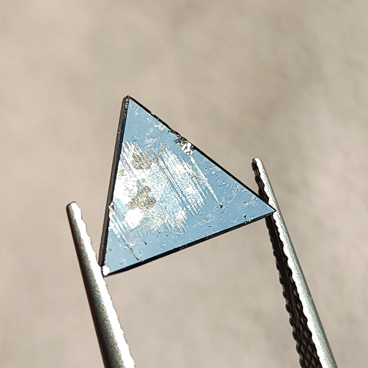 Flat back side of a triangular rose cut gemstone held in tweezers showing flat surface reflection.