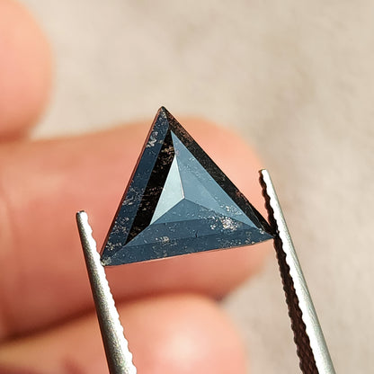 Triangular rose cut dark gemstone held in tweezers against a soft-focus background.