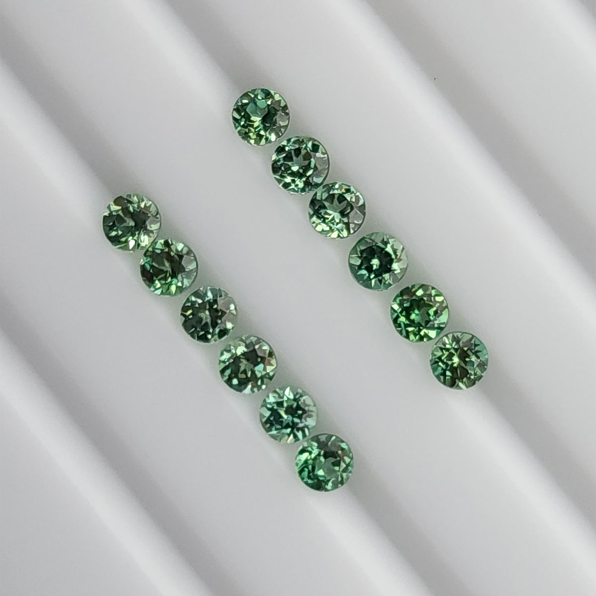 Mint-green round lab-grown sapphire stock arranged table-up in two rows on a white grooved tray.