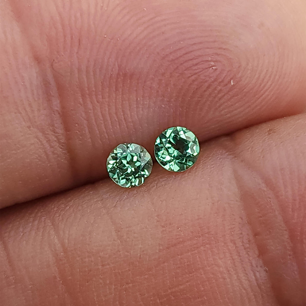 Two mint-green round lab-grown sapphires resting table-up on a fingertip for scale.