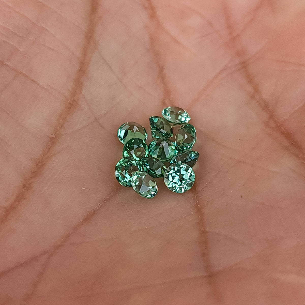 Group of mint-green round lab-grown sapphires resting in a palm for scale, with mixed facet orientations; sold per stone.