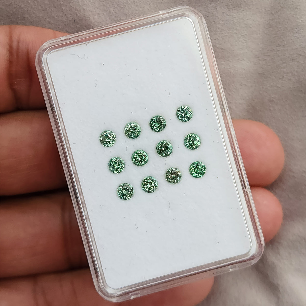 Hand holding a clear case of representative mint-green round sapphire stock arranged face-up.
