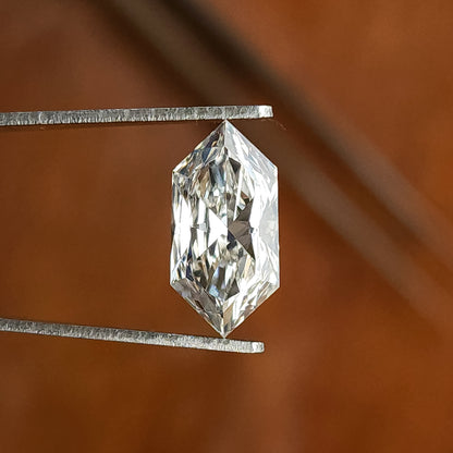 Elongated hexagonal cut colorless stone held vertically in metal tweezers against a brown backdrop.