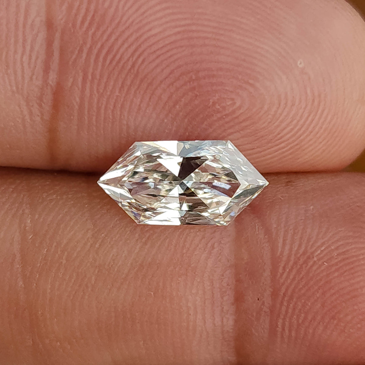 Face-up horizontal view of an elongated hexagon cut clear gem resting across human fingertips.