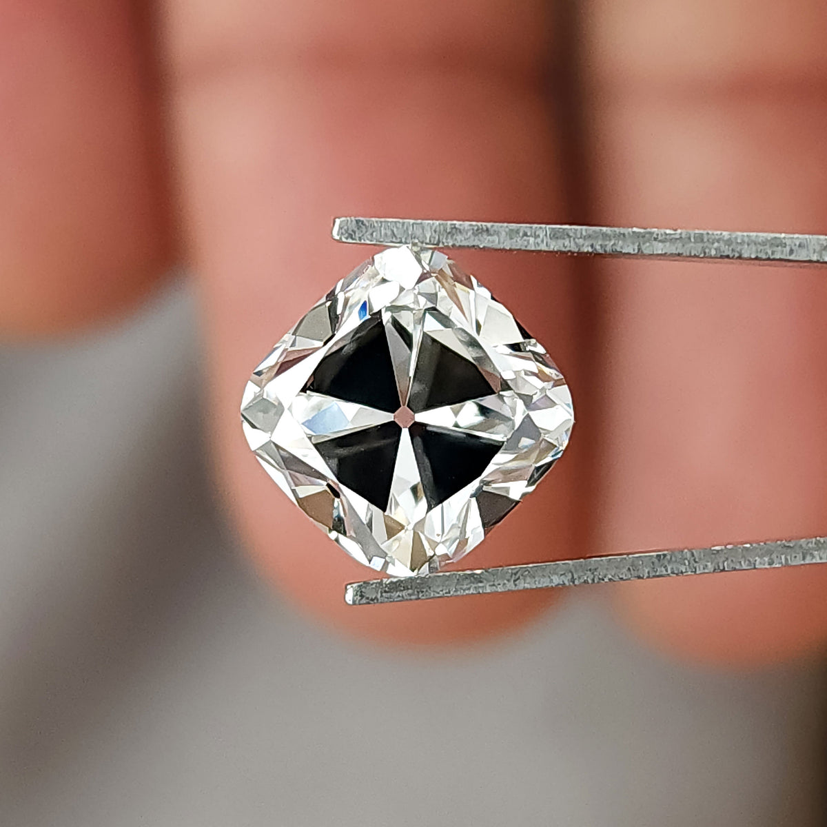 Square cushion cut colorless stone held in tweezers over fingers, showing table and facet reflections.