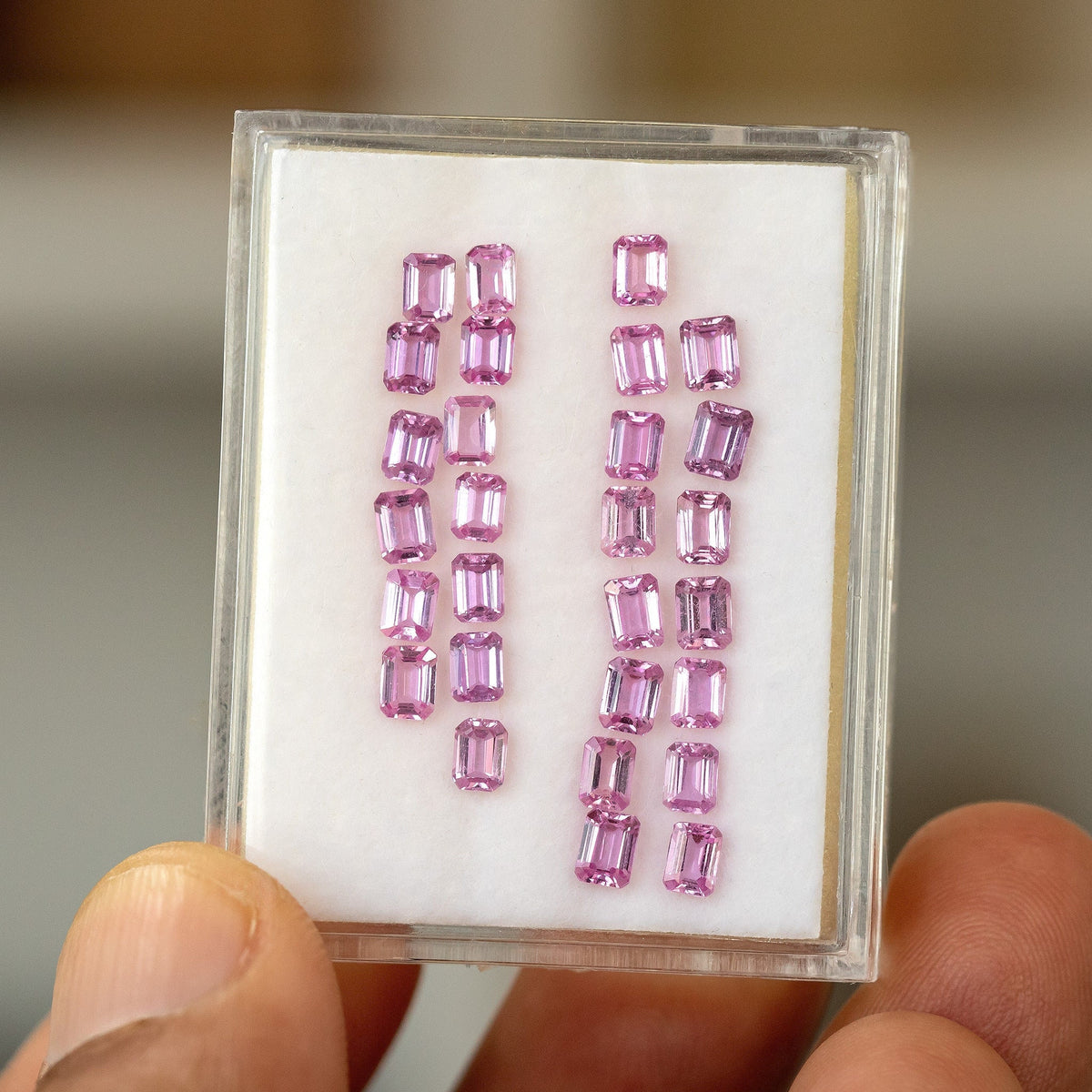 Representative parcel of pink emerald-cut sapphires in rows in a clear display tray held by fingers; price is for one randomly selected stone.