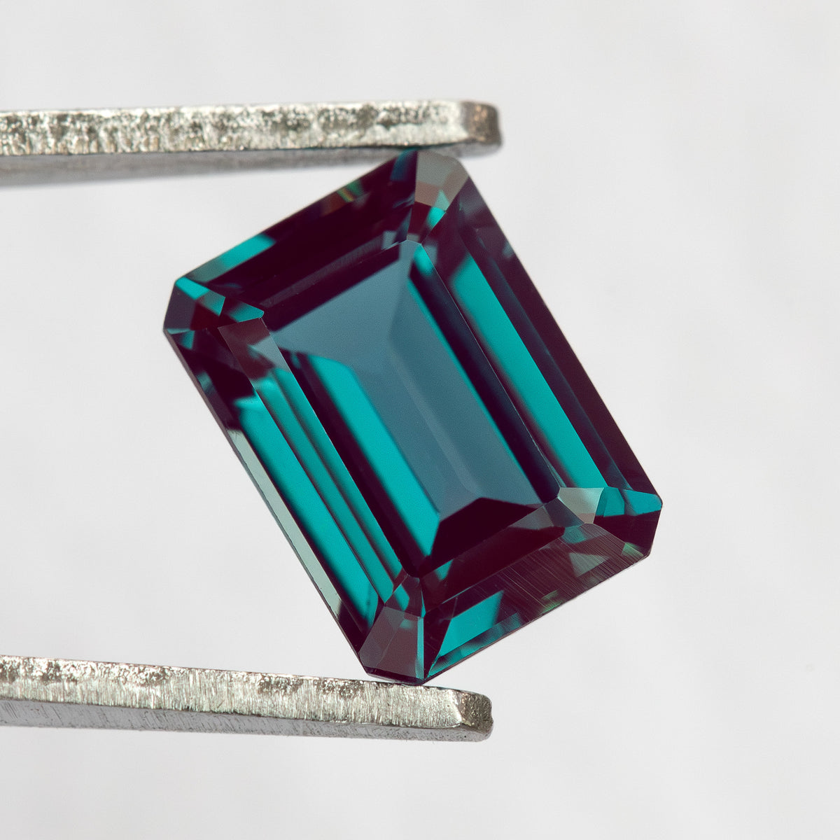 Emerald-cut lab-grown alexandrite sample held at an angle in tweezers, showing face-up step facets.