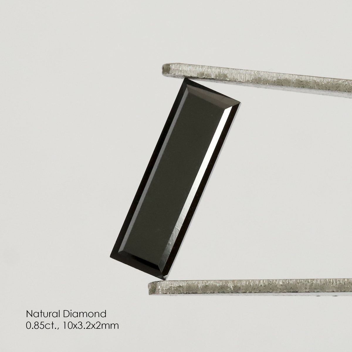 Reference black baguette diamond held diagonally in tweezers, with its complete rectangular outline and a bright reflection along one side.