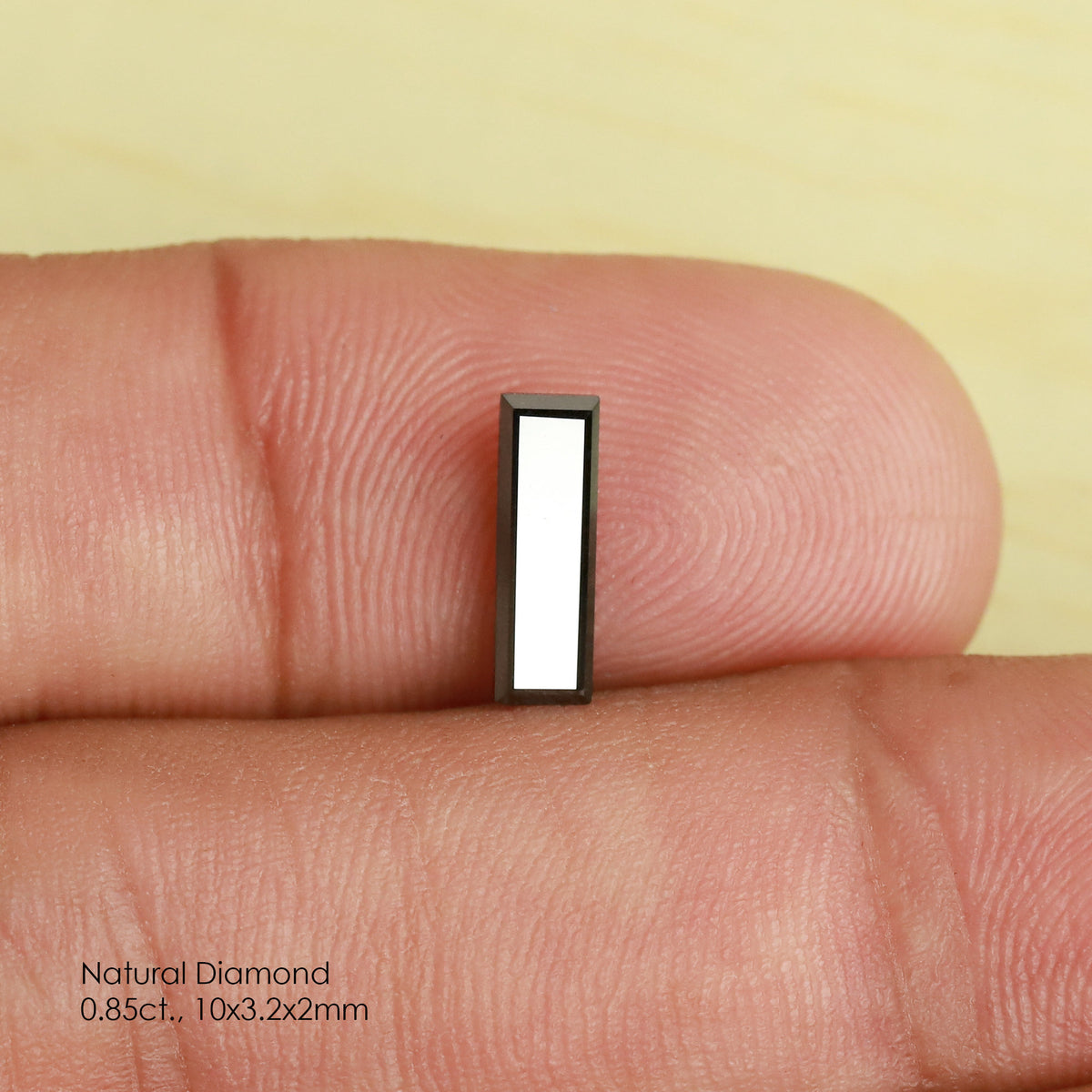 Reference black baguette diamond standing between fingertips for scale, with a broad white reflection across its table.
