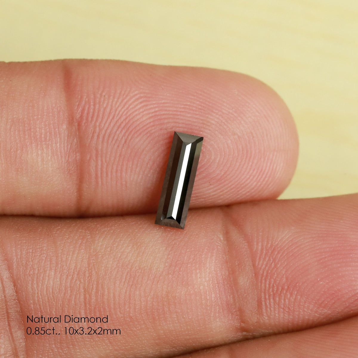Reference black baguette diamond tilted between fingertips, showing narrow bright facet reflections and its elongated outline.