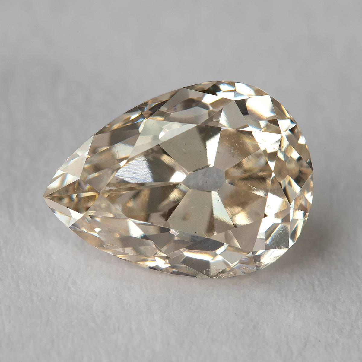 Face-up view of the exact light-champagne old-mine pear-cut diamond on a light surface, showing its outline, broad facets and visible characteristics.
