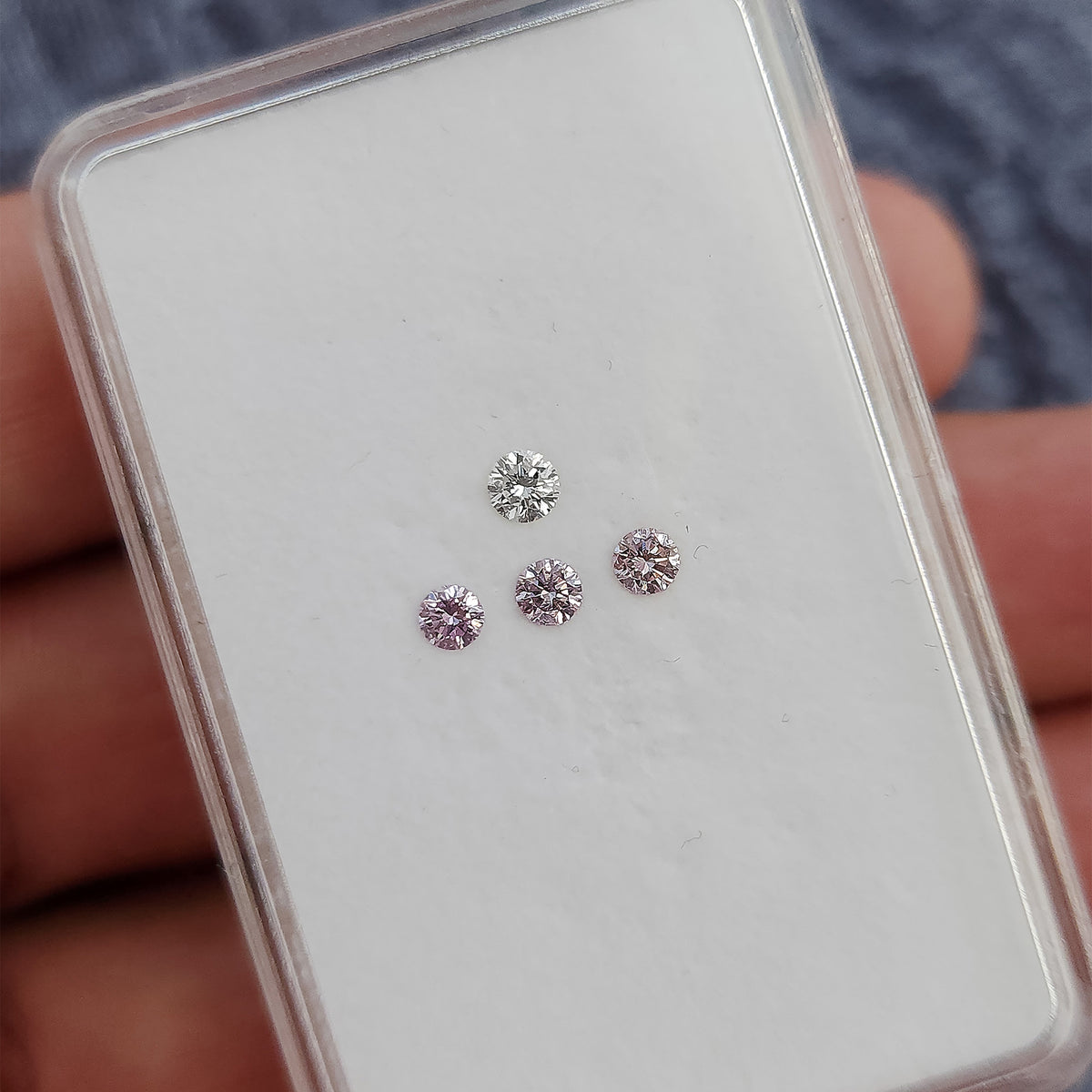 Tilted gem box held in a hand, showing pink round diamond samples with one white color reference.
