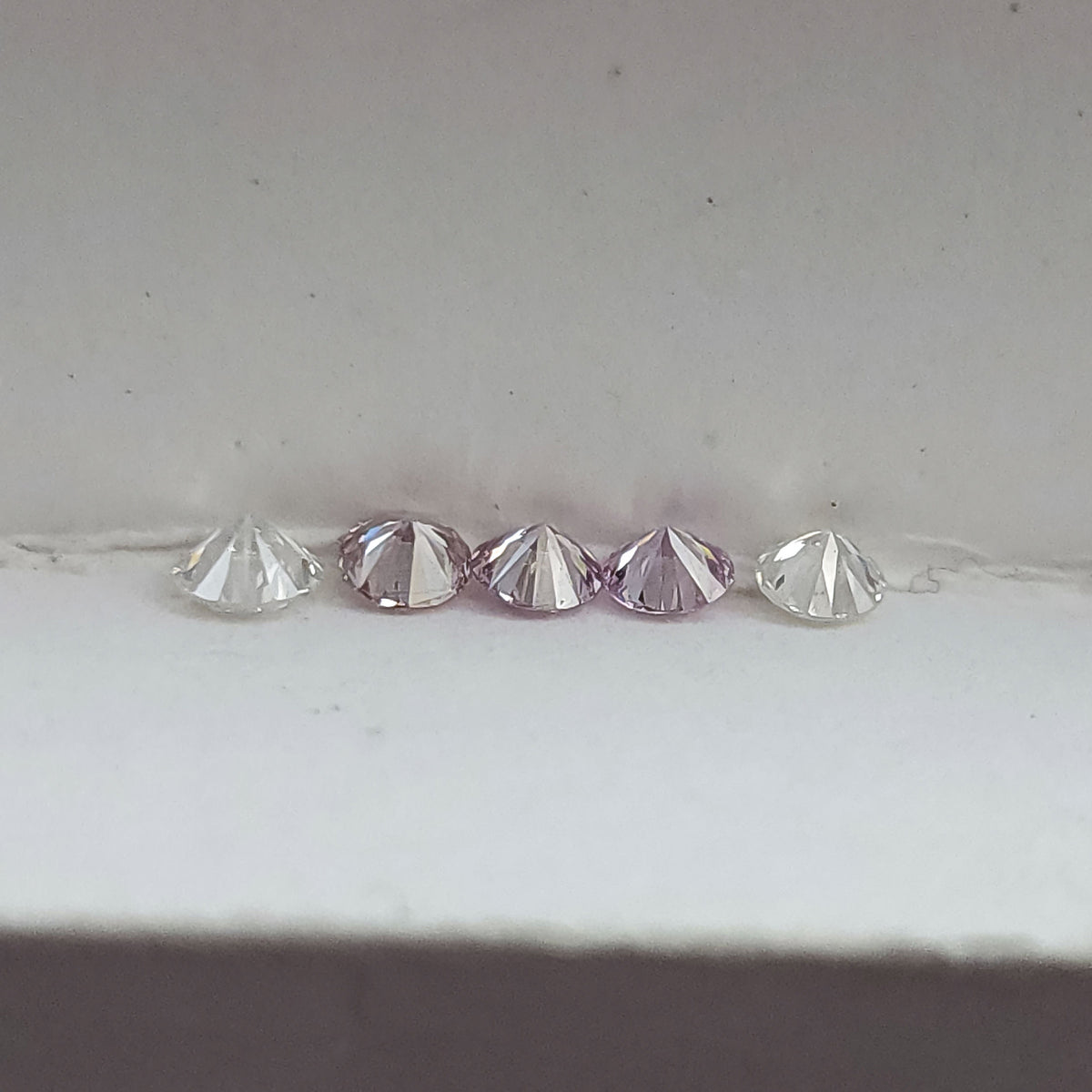 Close low-profile row of pink round diamond samples and white comparison stones, showing the pointed pavilion shapes.