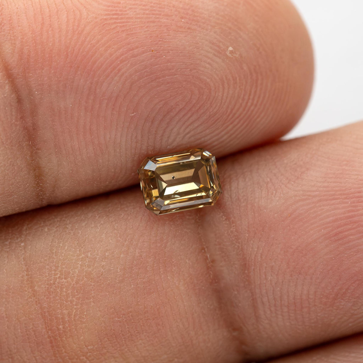 Face-up fingertip view of the 1.01 ct natural fancy brownish-yellow emerald cut diamond showing physical scale, warm golden-brown color and its visible dark clarity characteristic.