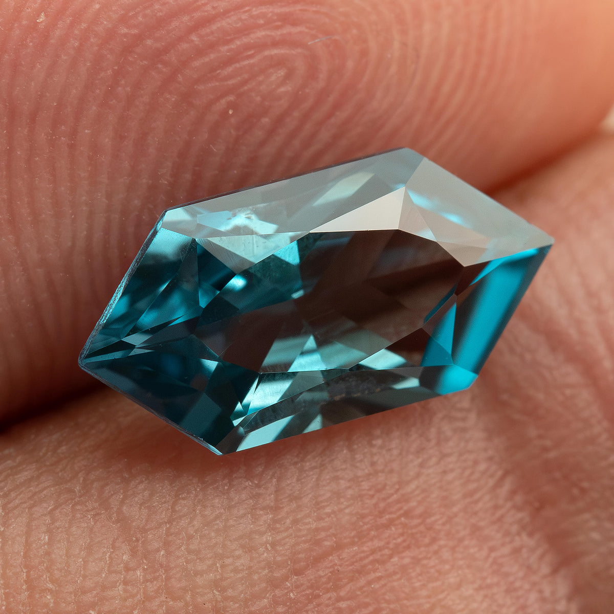 Teal lab-grown sapphire resting against fingers, showing its elongated shape and bright blue-green facets.