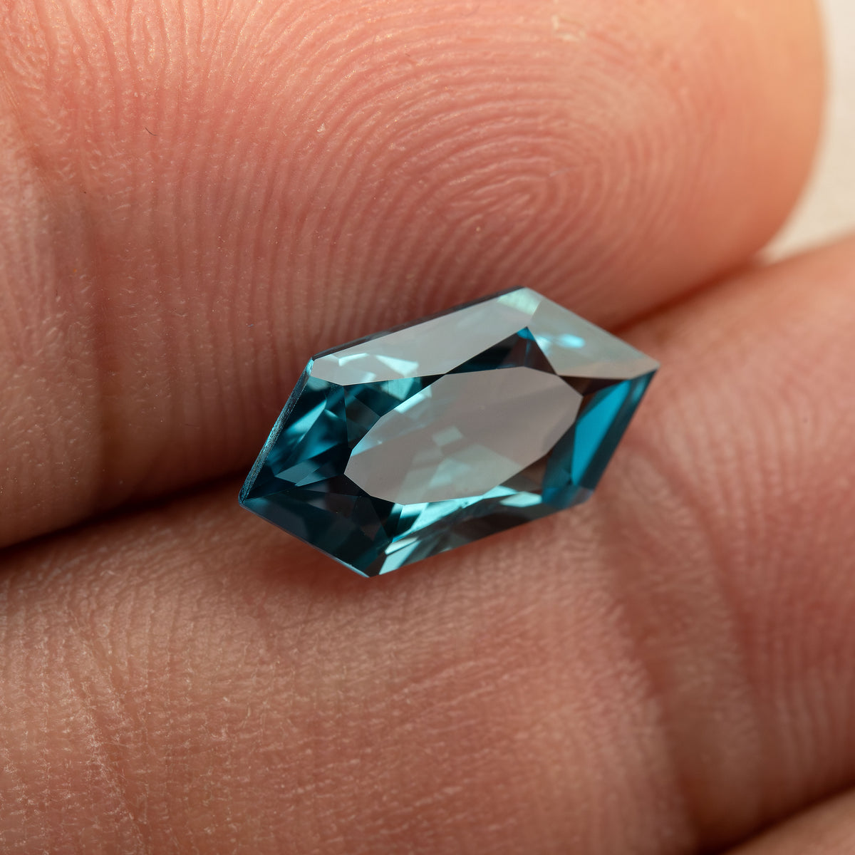 Teal lab-grown sapphire against a fingertip, showing its pointed ends and blue-green edge reflections.