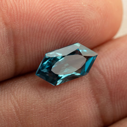 Teal lab-grown sapphire against a fingertip, showing its pointed ends and blue-green edge reflections.