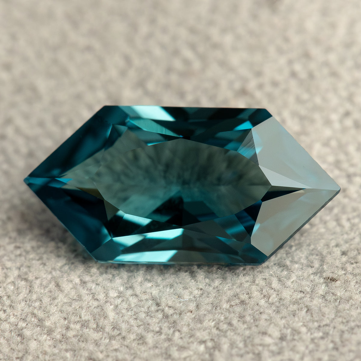 Horizontal face-up view of a teal lab-grown sapphire on a textured pale surface, with dark teal facet reflections.
