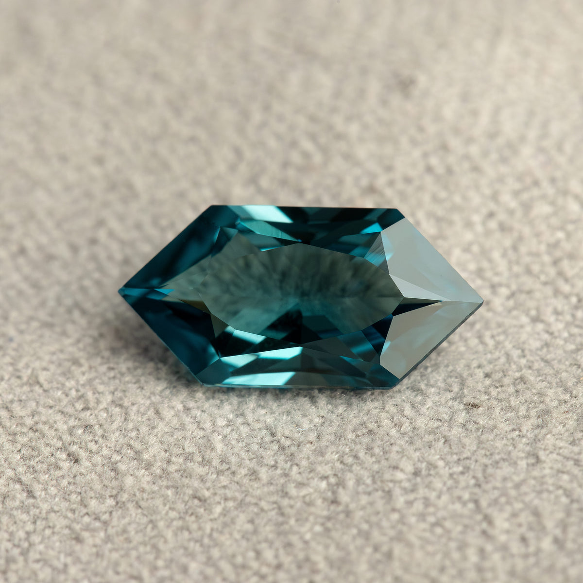 Wider view of a teal lab-grown sapphire on a textured pale background, showing its elongated hexagonal outline.