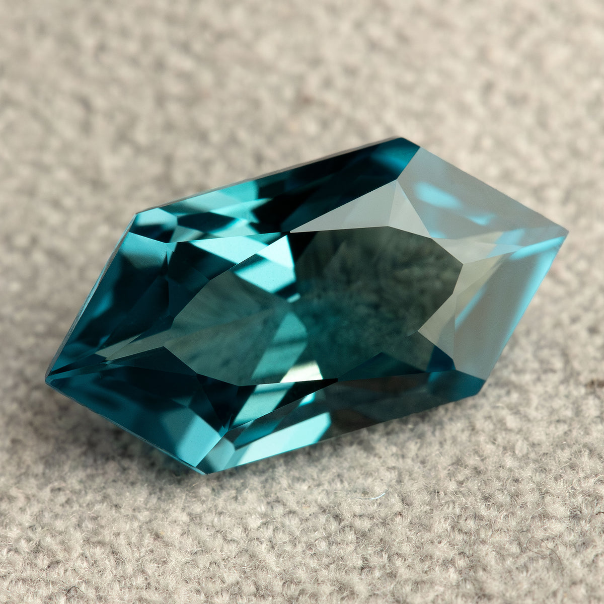 Angled close-up of a teal lab-grown sapphire on a textured pale surface, showing light and dark blue-green facets.