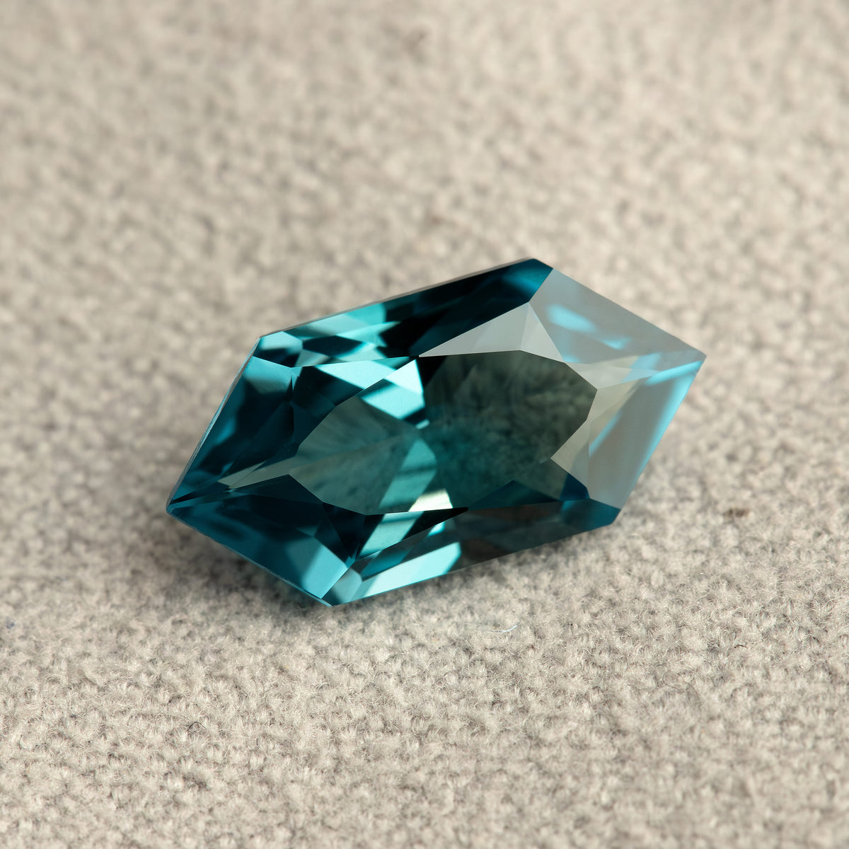 Angled teal lab-grown sapphire on a textured pale surface, showing bright blue-green and dark facet reflections.