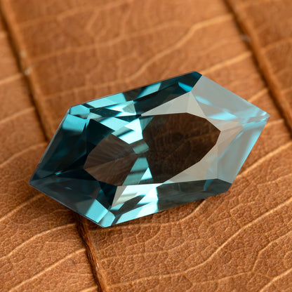 Teal lab-grown sapphire on a brown leaf, showing a broad central reflection and blue-green edge facets.