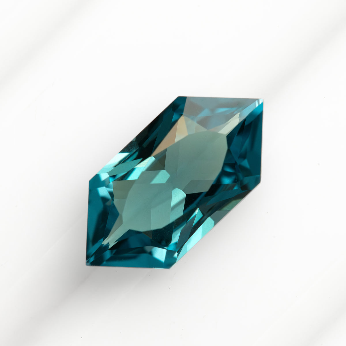 Teal lab-grown sapphire positioned diagonally on a white background, showing bright and dark blue-green facets.