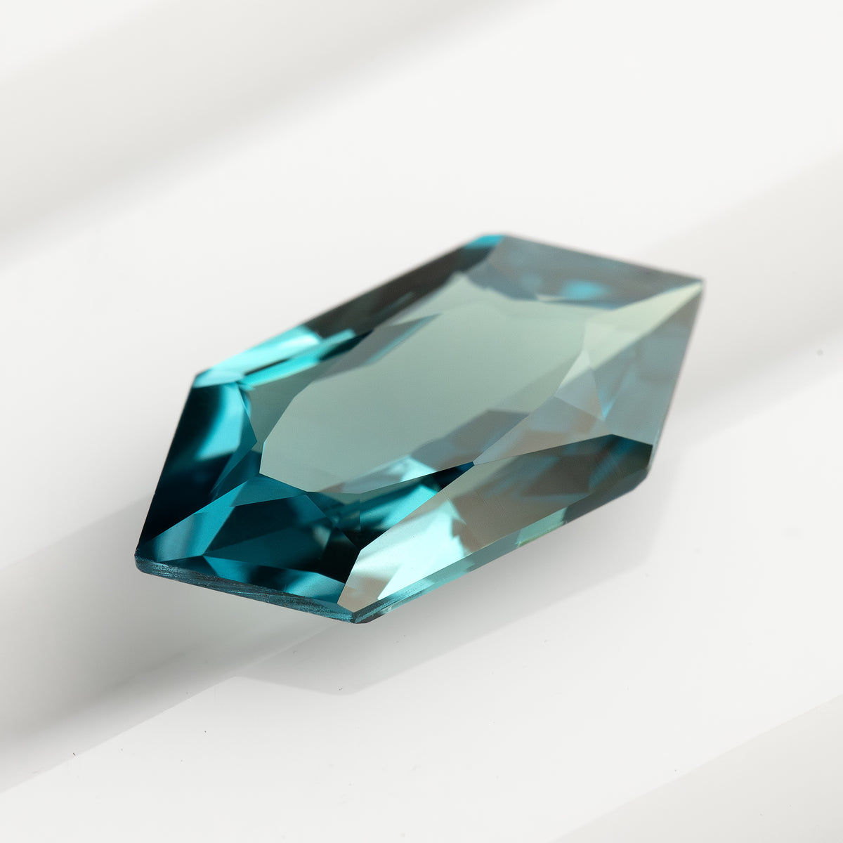 Angled teal lab-grown sapphire on a white background, showing a broad pale central reflection and blue-green edges.