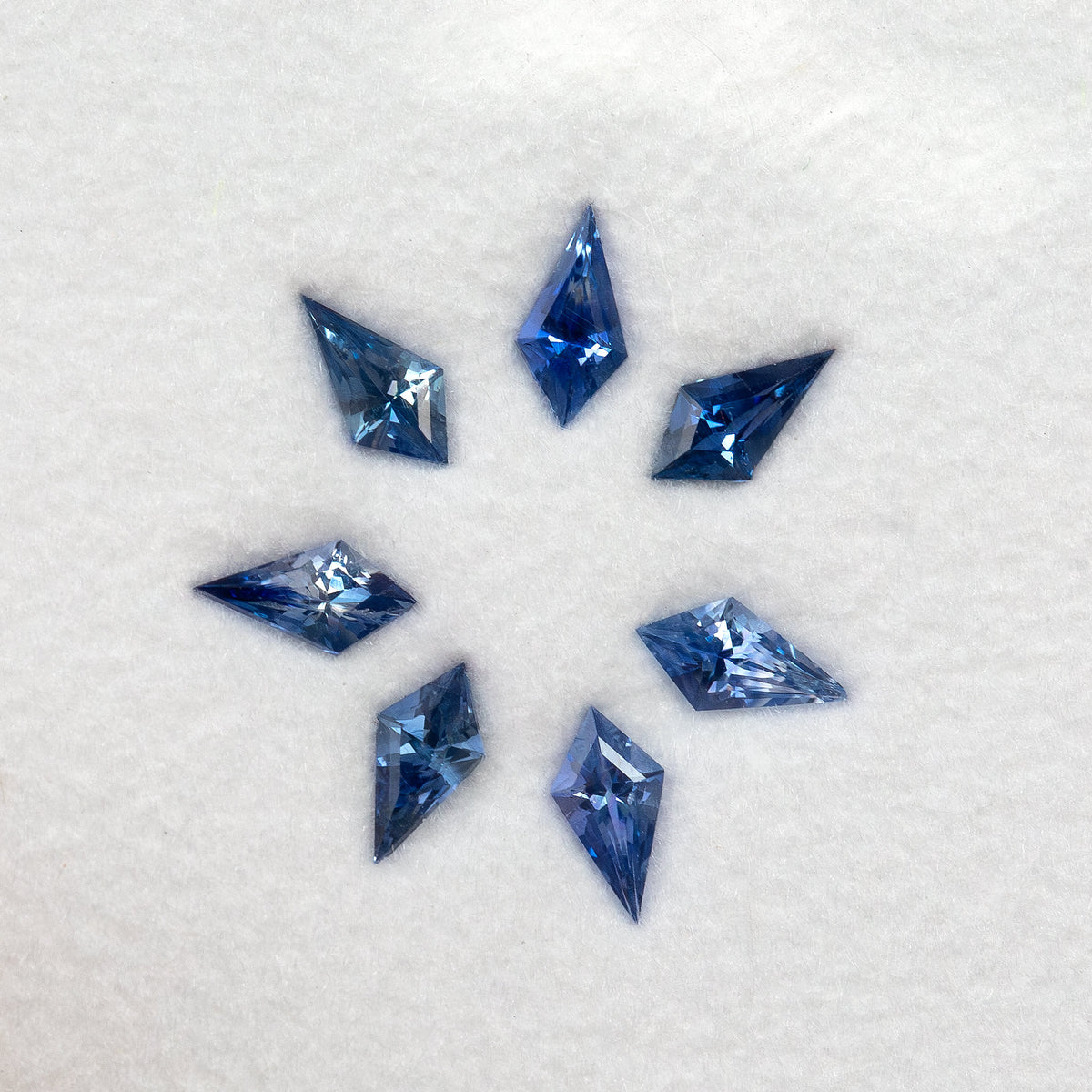 Seven blue kite-shaped sapphires arranged in a circle on a white pad, seen from above.