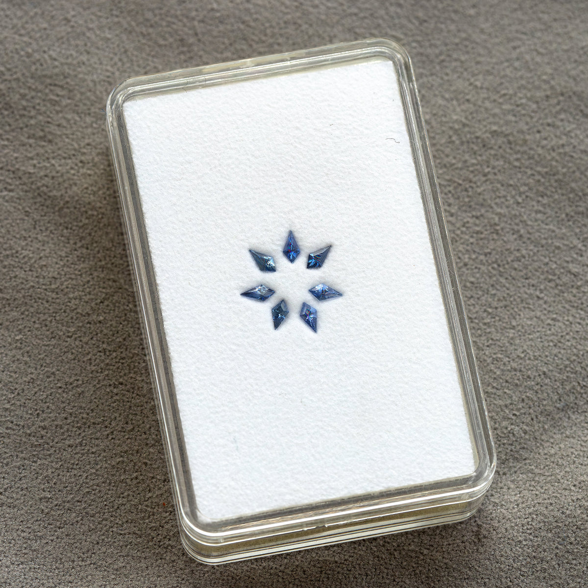 A complete rectangular gem box holding the seven blue kite-shaped sapphires on a white pad.