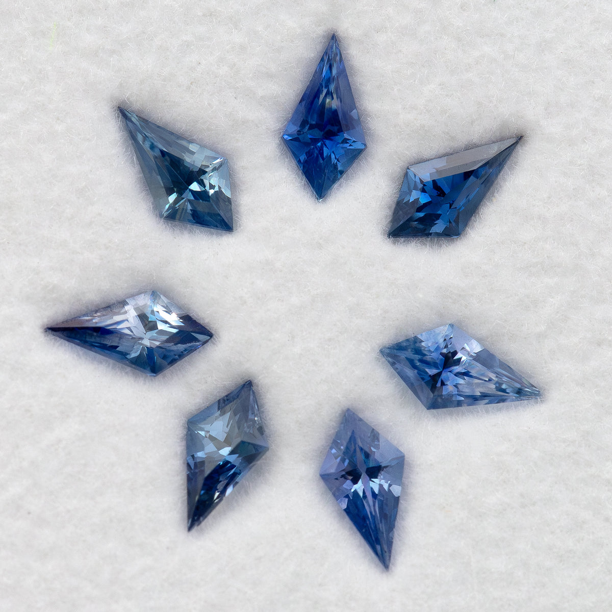Seven blue sapphires on a white pad, with pale and deeper blue reflections across the kite-shaped outlines.