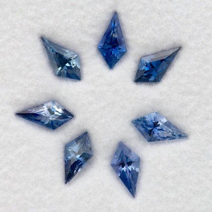 Seven blue sapphires on a white pad, with pale and deeper blue reflections across the kite-shaped outlines.