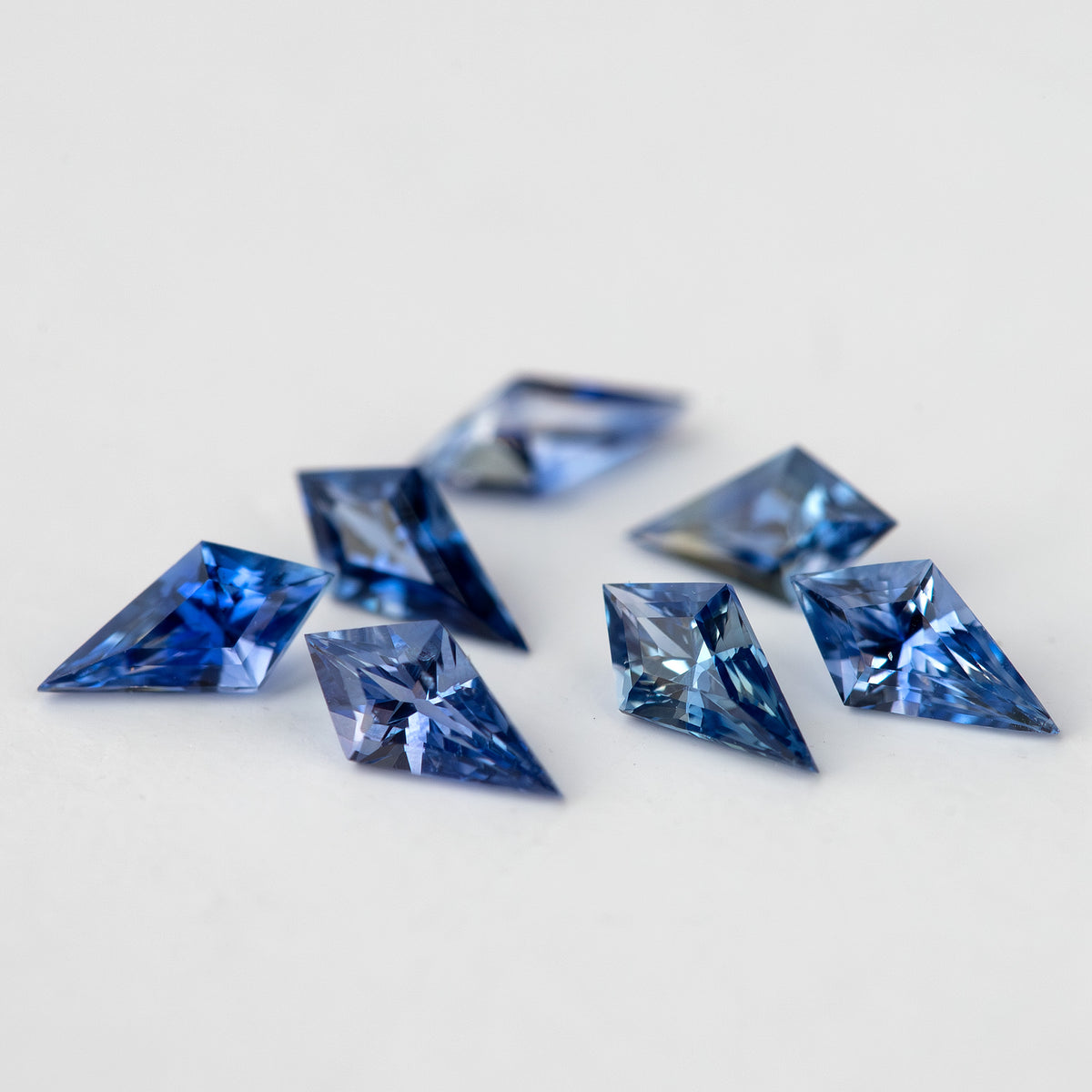 Seven loose blue sapphires photographed obliquely on white, with the front outlines and side facets visible.