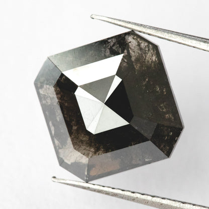 Brownish-black natural diamond held face-up in tweezers, showing the clipped-square outline, broad triangular facets and visible internal markings.