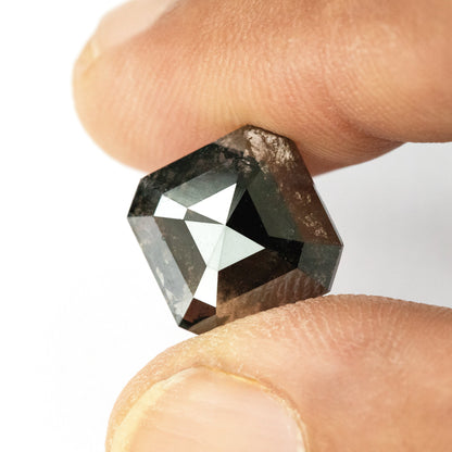 Brownish-black diamond held between two fingertips, with the full face-up outline and mottled internal markings visible.