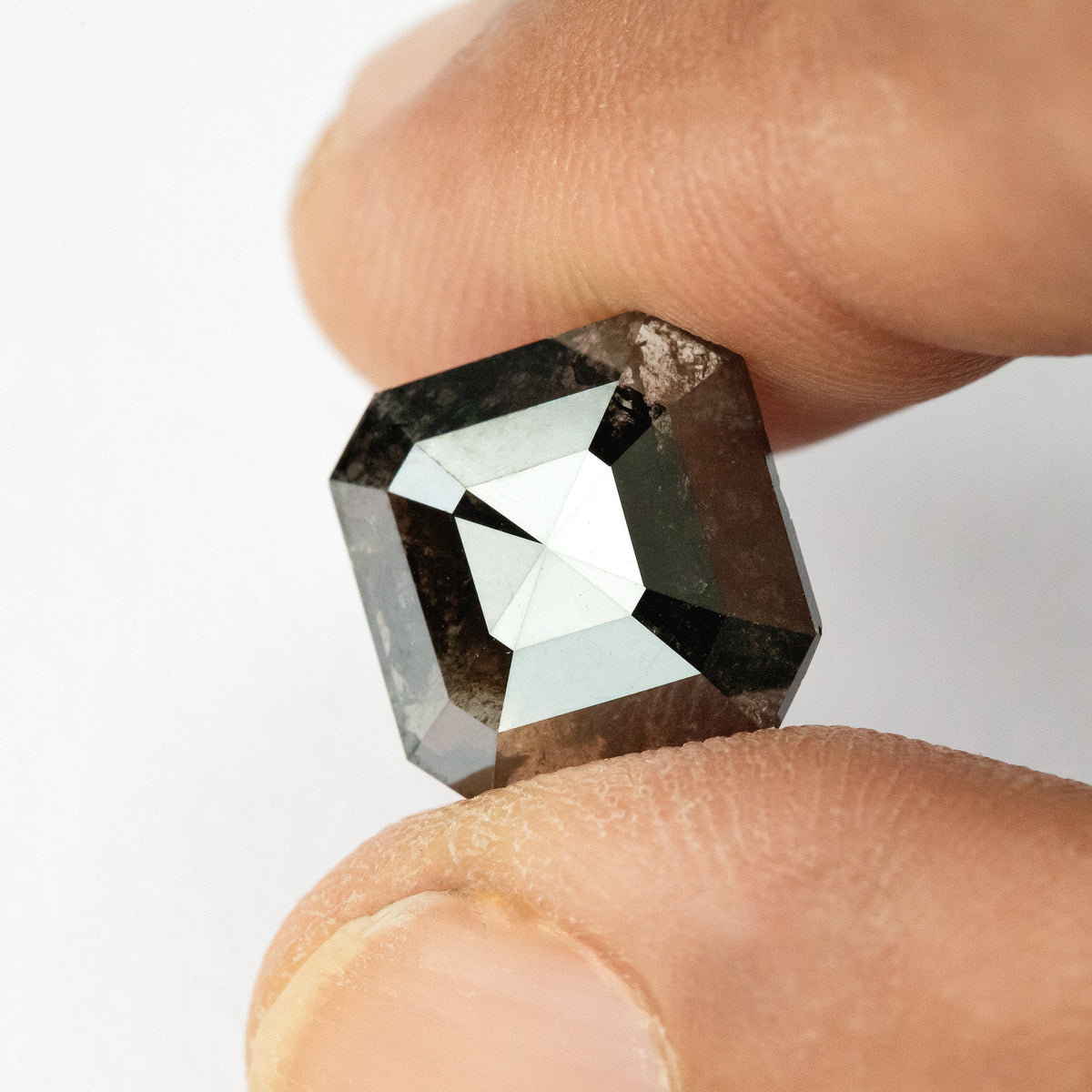 Alternate face-up view of the brownish-black diamond between fingertips, showing its clipped corners and dark triangular reflections.