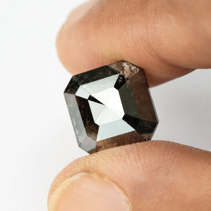 Alternate face-up view of the brownish-black diamond between fingertips, showing its clipped corners and dark triangular reflections.